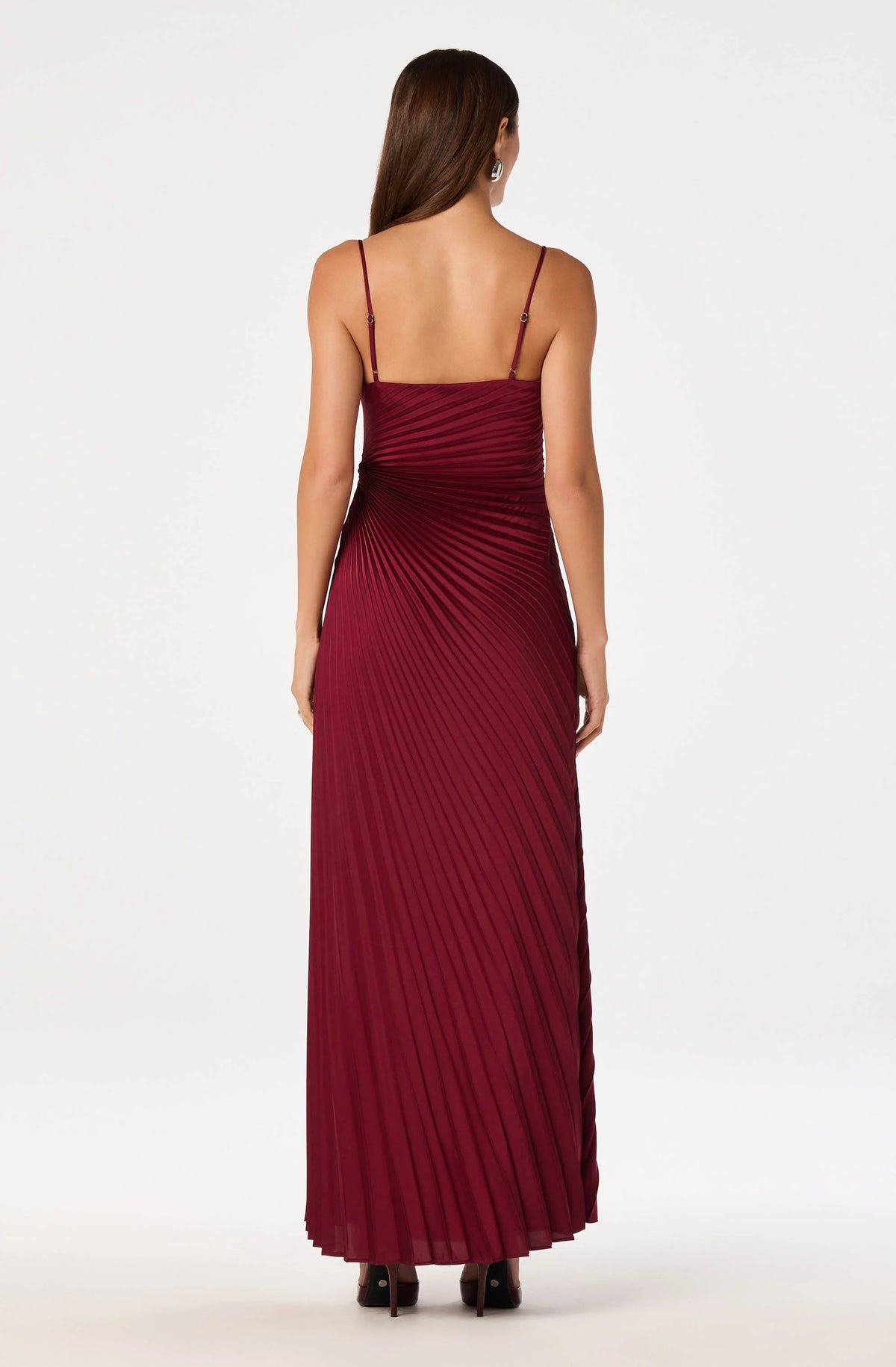 A woman is shown from the back wearing a rich burgundy satin maxi dress with fine sunburst pleating and adjustable spaghetti straps.