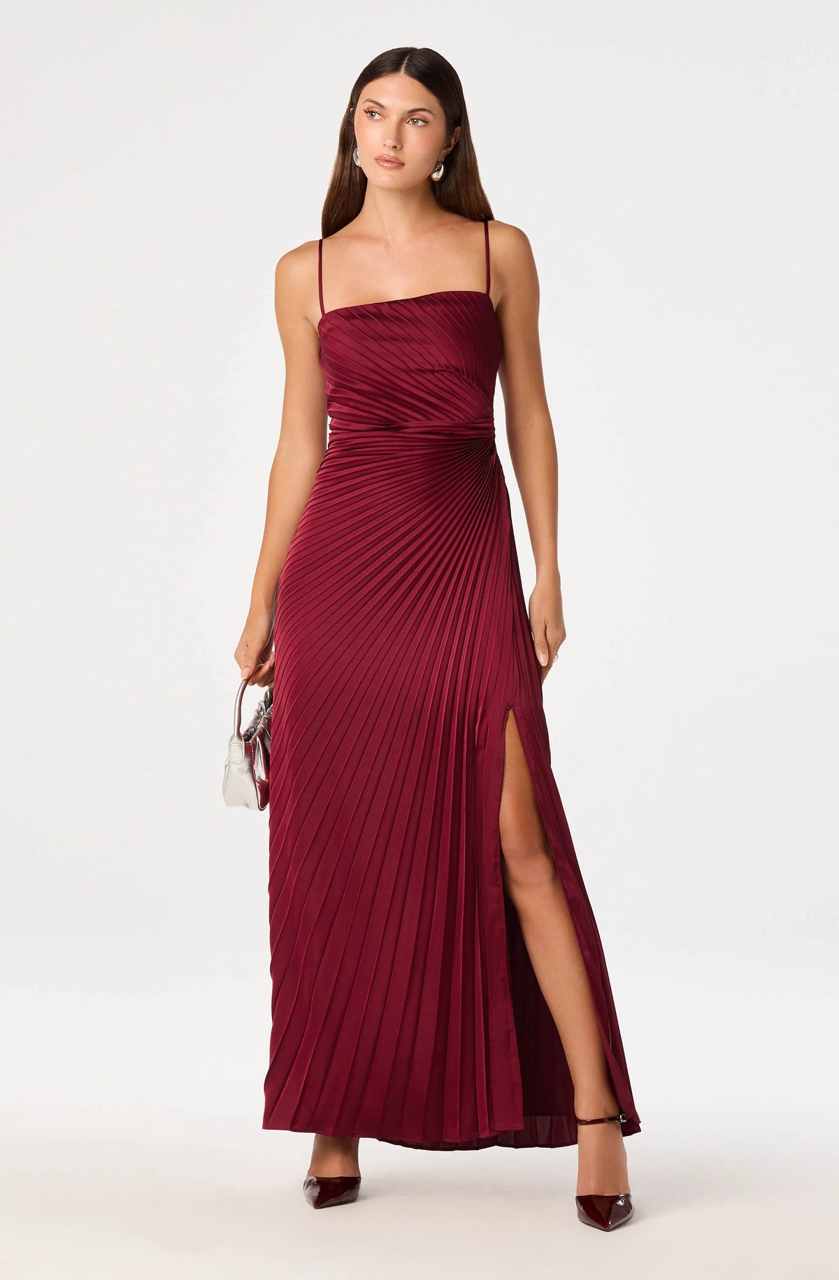 A model showcases a stunning burgundy satin maxi dress featuring fine sunburst pleating and a leg slit, ideal for elegant events like galas or weddings. This fashionable dress is conveniently available near Apex, Cary, and Raleigh.