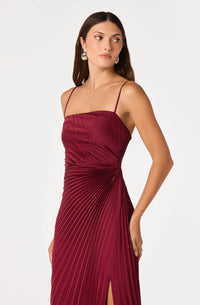 A model poses in a rich burgundy satin maxi dress featuring fine sunburst pleating and adjustable spaghetti straps, showcasing a side leg slit.
