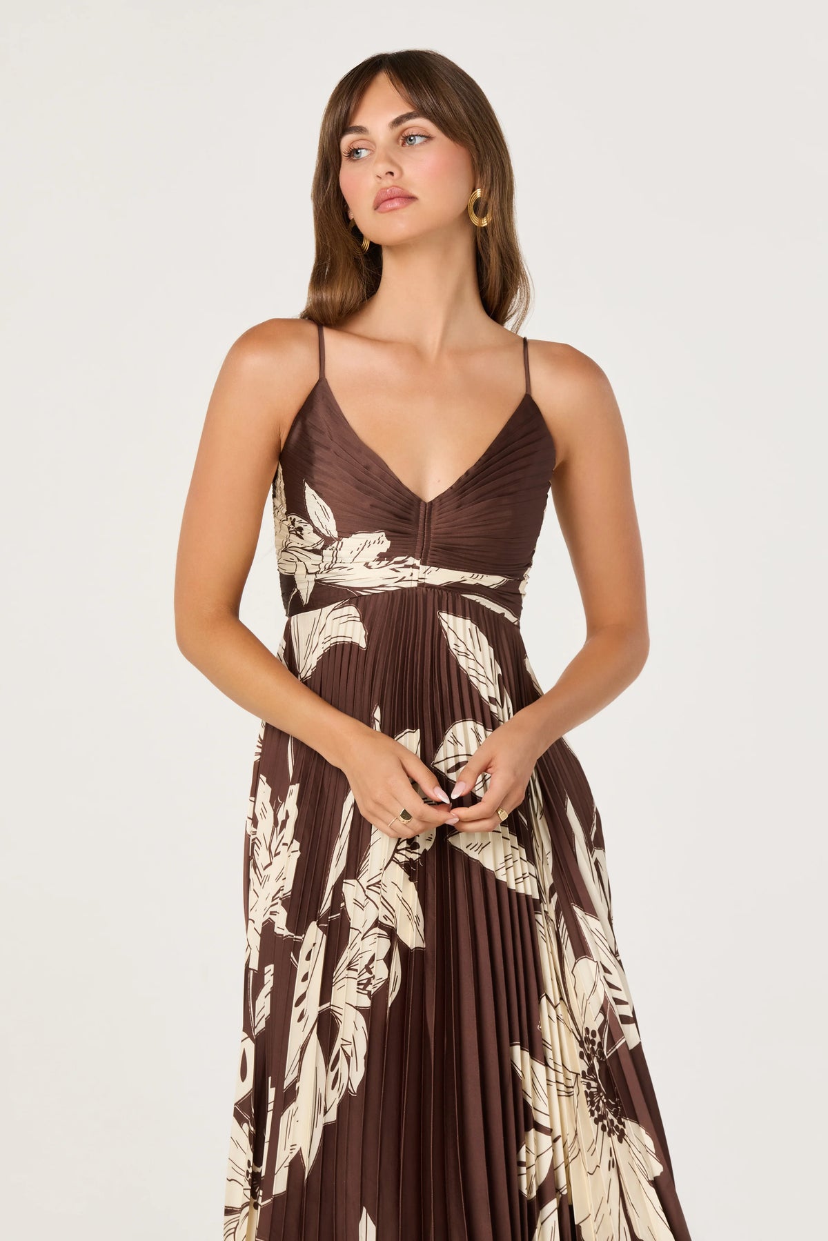 A model wearing a pleated satin dress with a deep V-neckline and adjustable spaghetti straps, featuring a floral print in shades of brown and cream.