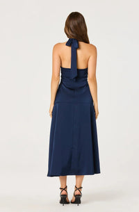 A woman is wearing a navy blue sleeveless dress with a halter neckline tied at the back, showcasing a fitted bodice and a flowing midi skirt.