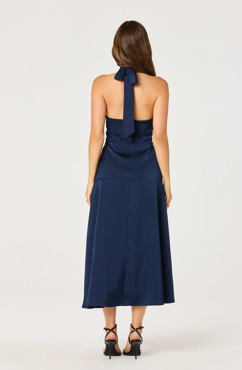 A woman stands with her back facing the camera, showcasing a navy blue satin halter dress with a bow tie at the neck. This elegant dress, perfect for events like a wedding or gala, is conveniently available near Apex, Cary, and Raleigh.