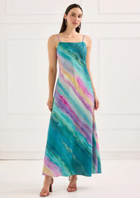 A model stands in a brightly lit room wearing a long, colorful dress featuring swirling patterns in shades of teal, pink, and purple.