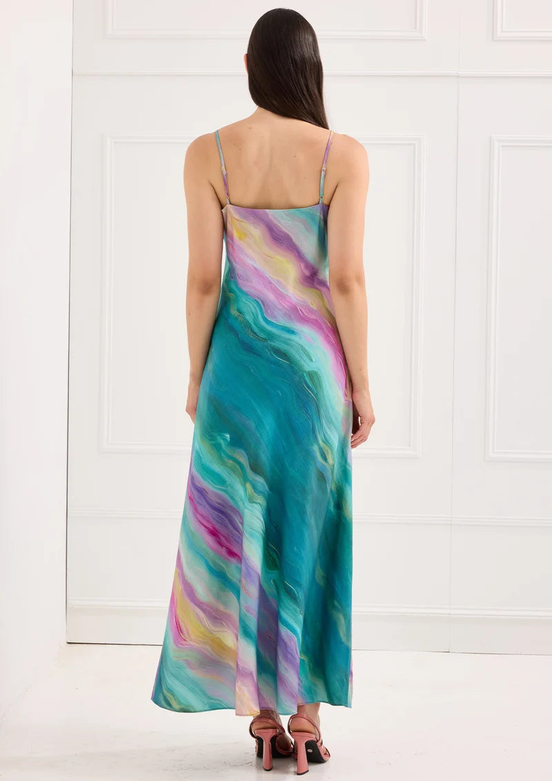 A model stands with her back to the camera, showcasing a flowing, multicolored maxi dress with adjustable straps.