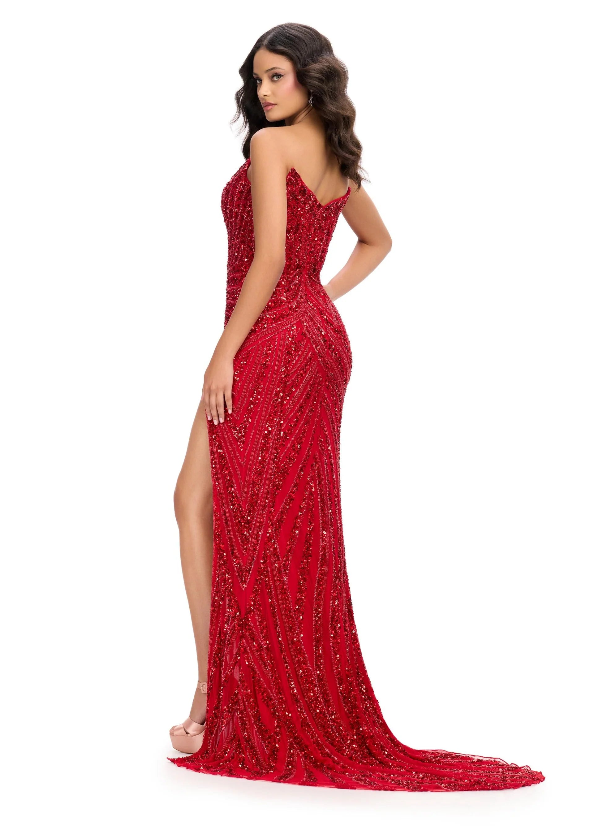 A model poses in a stunning red strapless gown adorned with intricate beadwork and a thigh-high slit. Perfect for a gala, prom, or black tie wedding, this elegant dress is conveniently available near Apex, Cary, and Raleigh.