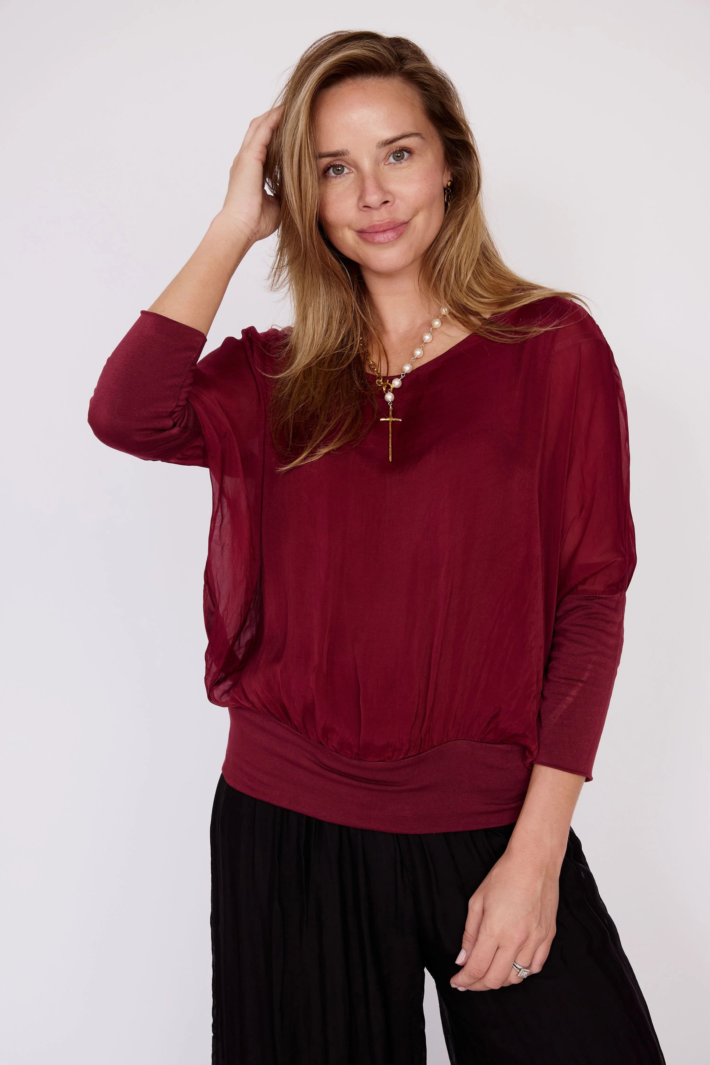 A woman stands confidently in a rich wine-colored silk top, featuring a loose georgette overlay and a banded hemline.