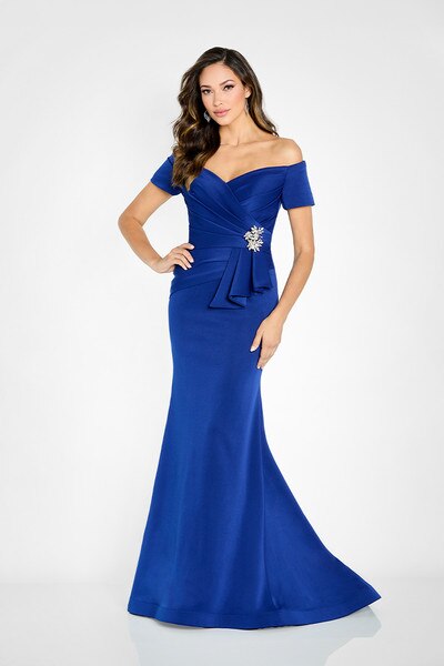 A model showcases a stunning sapphire blue trumpet gown with off-the-shoulder sleeves and a decorative brooch at the waist.