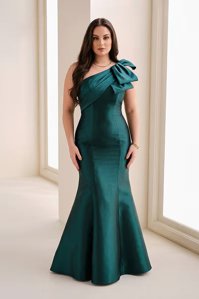 A woman models a stunning emerald green fit and flare gown with a chic one-shoulder neckline and a dramatic bow detail.