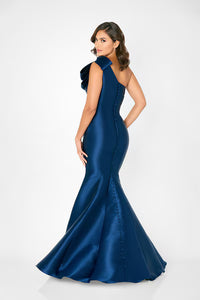 A model poses in a stunning navy blue mermaid gown featuring a dramatic ruffled shoulder and a fitted silhouette with button detailing along the back. This elegant dress is perfect for formal events such as black tie weddings or galas.