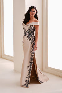 A model stands elegantly in a champagne-colored trumpet gown with intricate black floral embroidery, showcasing an off-the-shoulder neckline and a side slit.