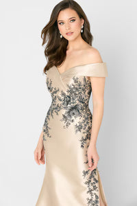 A model showcases an elegant champagne trumpet gown with off-the-shoulder sleeves and intricate black floral embroidery.