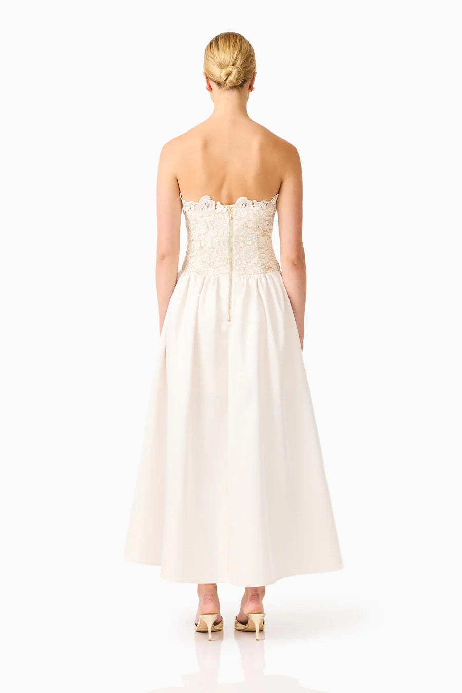 A model is seen from the back wearing a strapless ivory gown featuring intricate lace detailing on the bodice and a flowy skirt.