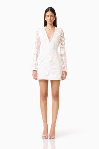 A model showcases a stylish white mini dress featuring long sleeves and a deep V-neckline, adorned with intricate 3D floral details.