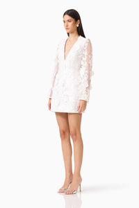 A model poses in a white mini dress featuring long flared sleeves and a deep V-neckline, adorned with 3D floral details.