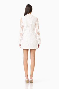 A model is shown from the back wearing a white mini dress with flared sleeves made of textured 3D floral fabric.