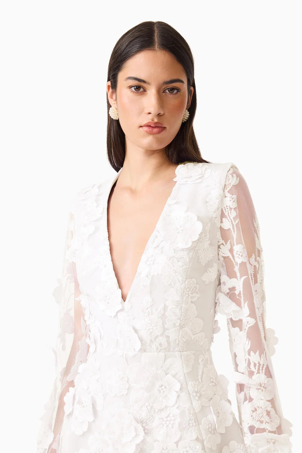 A model showcases a stunning white mini dress featuring a deep V-neckline and elegant flared sleeves adorned with intricate 3D floral details.
