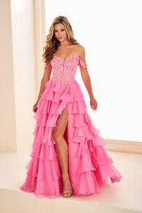 A model poses in a vibrant hot pink off-the-shoulder gown featuring three-dimensional flower lace straps and a front slit, showcasing a layered ruffled skirt.