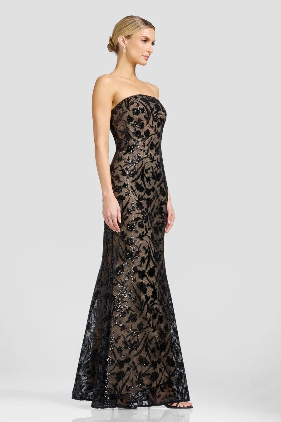 A model showcases a stunning strapless evening gown featuring intricate black floral patterns and shimmering embellishments.