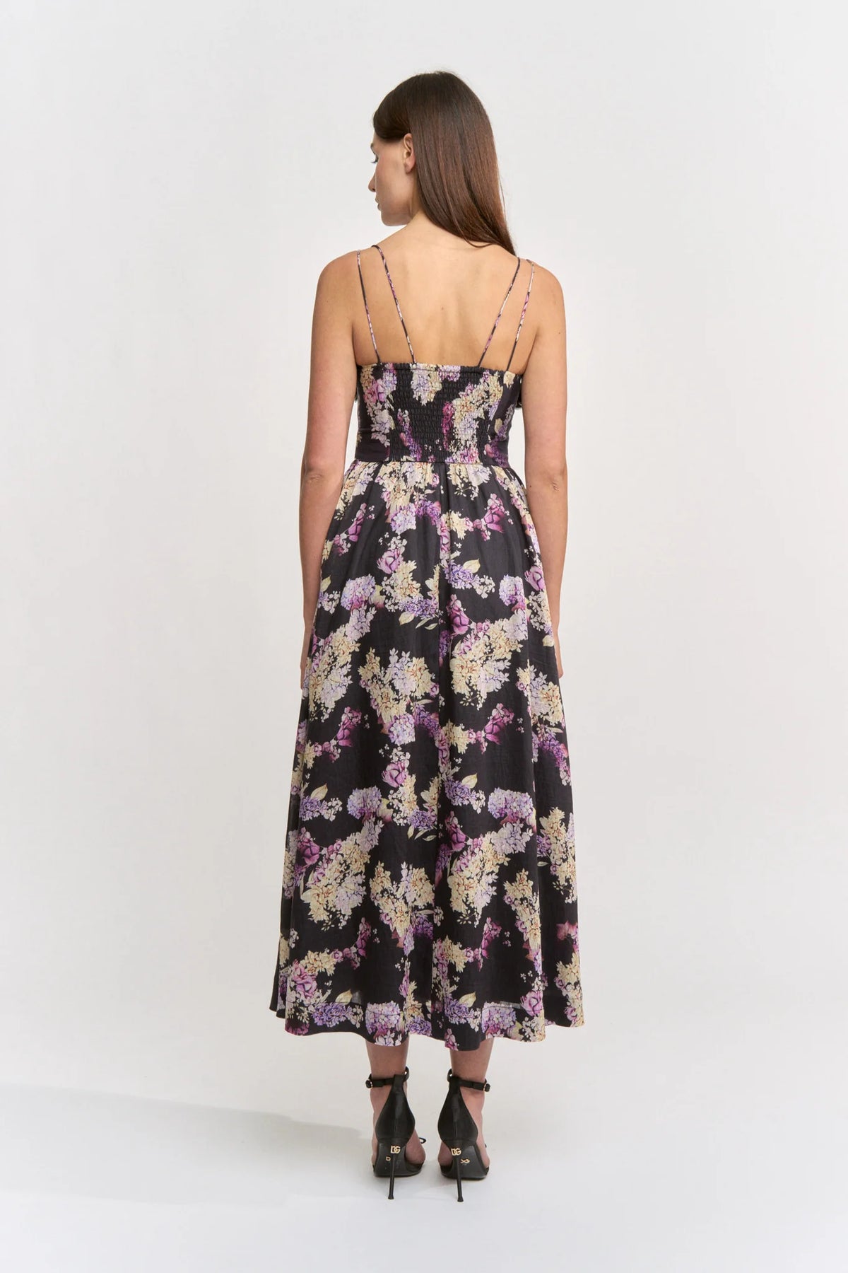 A model stands with her back to the camera, showcasing a sleeveless floral midi dress featuring a sweetheart neckline and delicate straps.