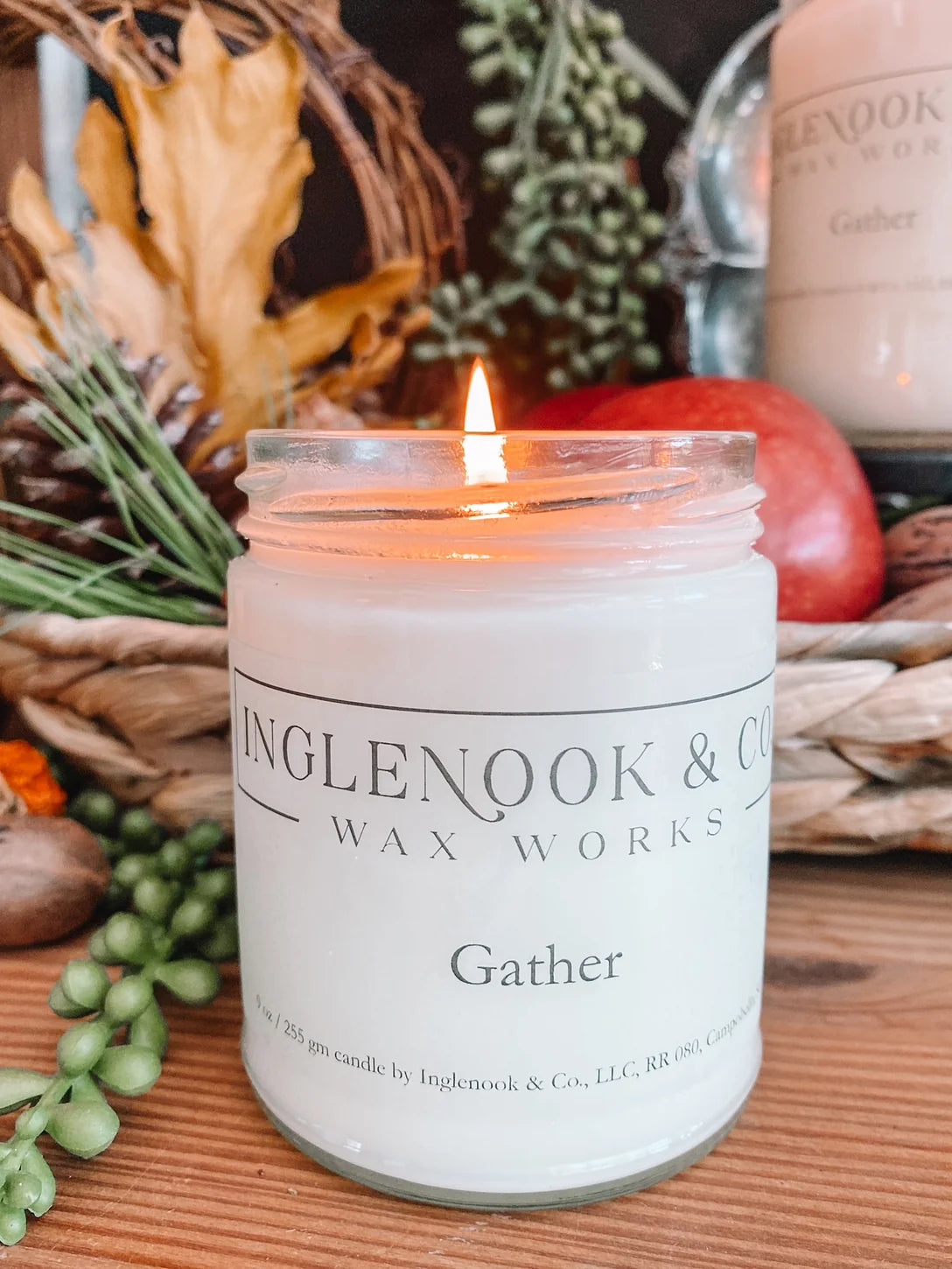 A lit Gather soy wax candle in a clear glass jar sits atop a wooden surface, surrounded by autumn leaves, apples, and greenery. The warm glow of the flame enhances the cozy atmosphere, perfect for gatherings during the fall season.
