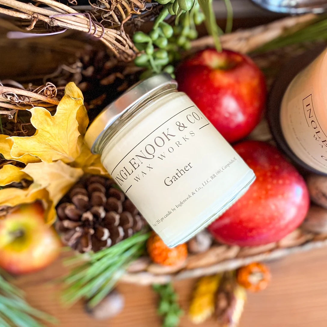 A Gather soy wax candle is displayed among autumn-themed decorations, including bright red apples, pine cones, and colorful leaves.