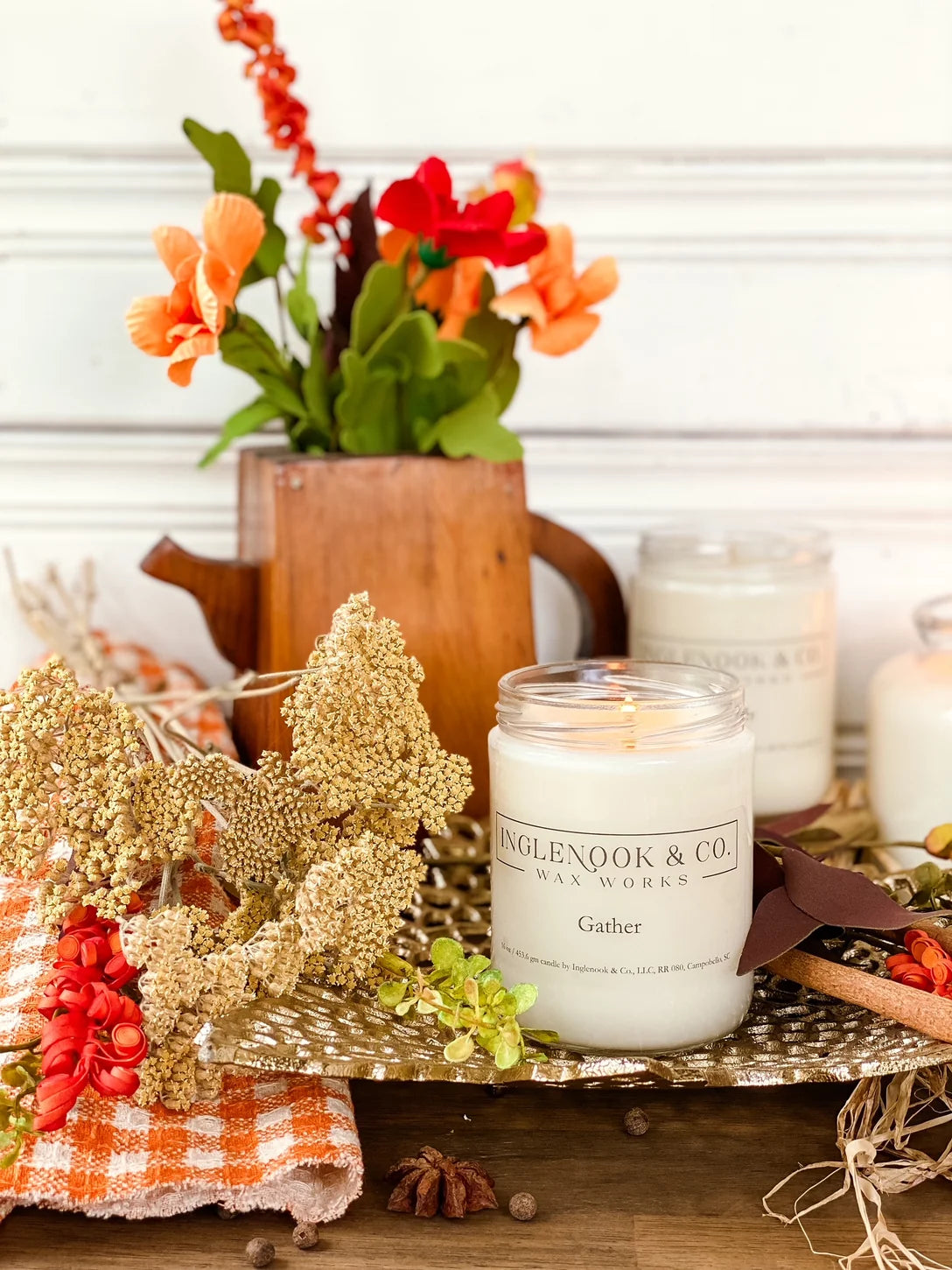A beautifully styled scene featuring the Gather Soy Wax Candle in a clear glass jar, surrounded by dried flowers, a wooden teapot, and decorative textiles. The warm ambiance is enhanced by the cozy, inviting colors of the arrangement.