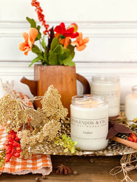 A beautifully styled scene featuring the Gather Soy Wax Candle in a clear glass jar, surrounded by dried flowers, a wooden teapot, and decorative textiles. The warm ambiance is enhanced by the cozy, inviting colors of the arrangement.