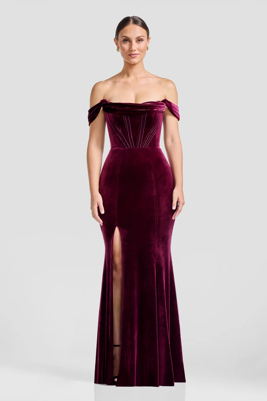 A model showcases a stunning off-the-shoulder gown in rich burgundy velvet, featuring a fitted bodice with corset boning and a dramatic mermaid silhouette with a thigh-high slit.