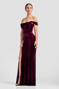 A model showcases a stunning off-the-shoulder gown in deep burgundy velvet, featuring a fitted bodice with exposed boning and a dramatic slit.