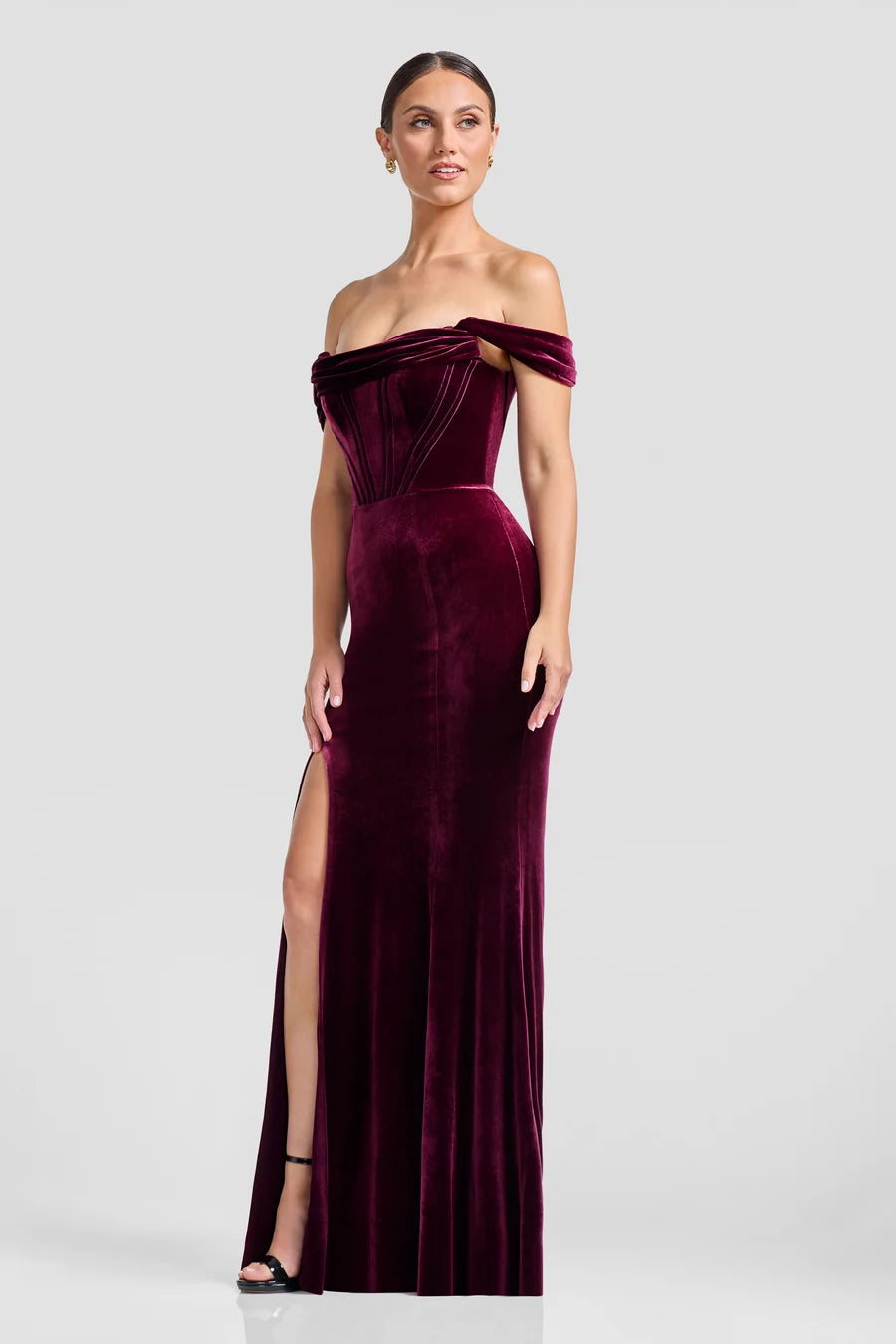 A model showcases a stunning off-the-shoulder gown in deep burgundy velvet, featuring a fitted bodice with exposed boning and a dramatic slit.