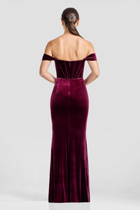 A woman models a stunning off-the-shoulder gown in rich burgundy velvet, showcasing a fitted mermaid silhouette with a subtle slit.