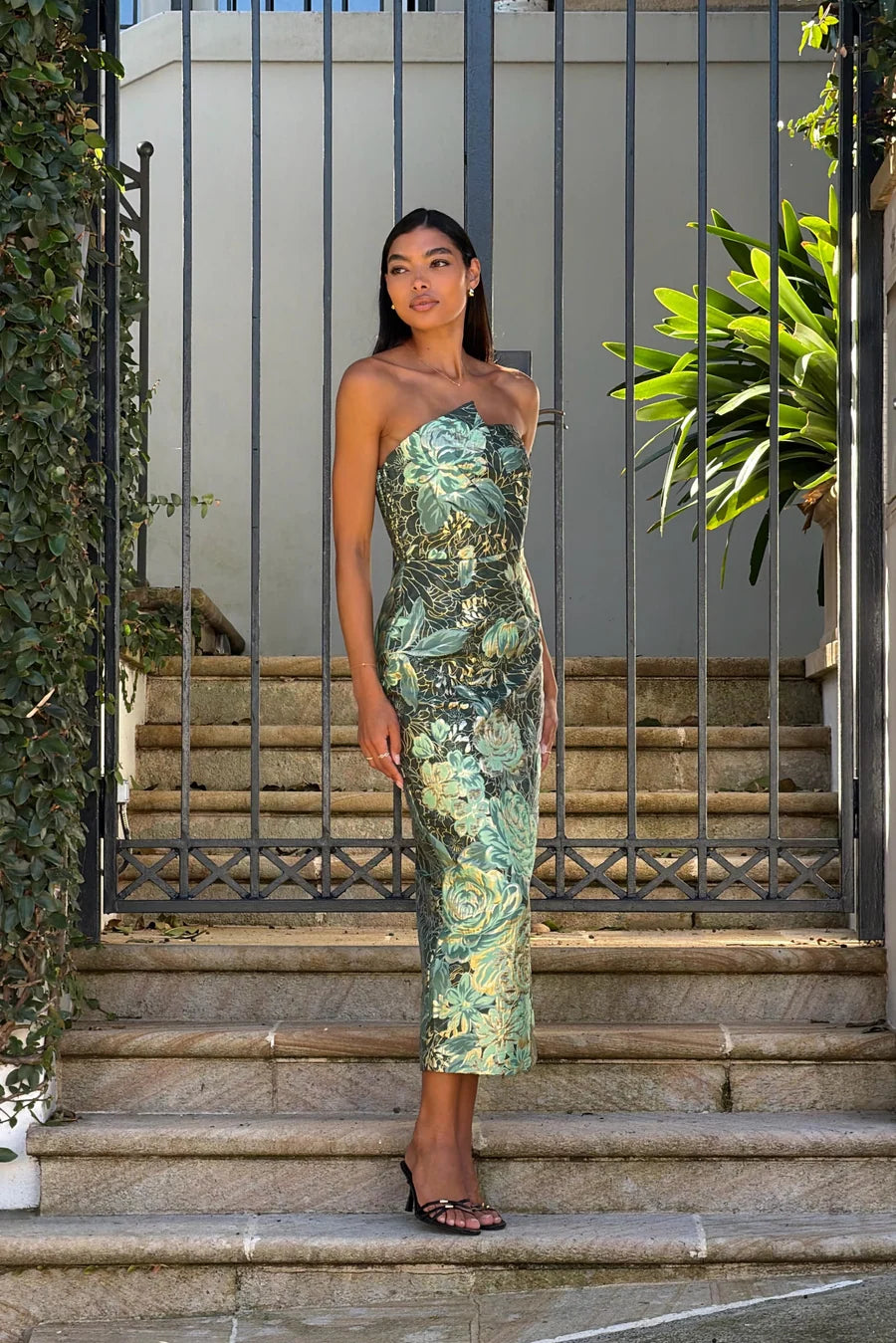 A model poses gracefully in a strapless, form-fitting green floral midi dress, standing on stone steps in front of a wrought iron gate surrounded by lush greenery.