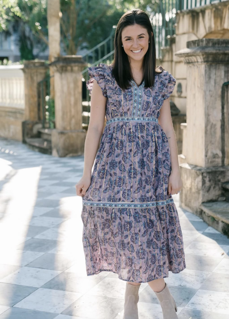 A model stands on a sunlit outdoor patio, wearing a flowing, floral-patterned dress with short flutter sleeves and a tiered skirt.
