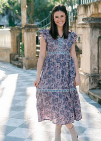 A model stands on a sunlit outdoor patio, wearing a flowing, floral-patterned dress with short flutter sleeves and a tiered skirt.