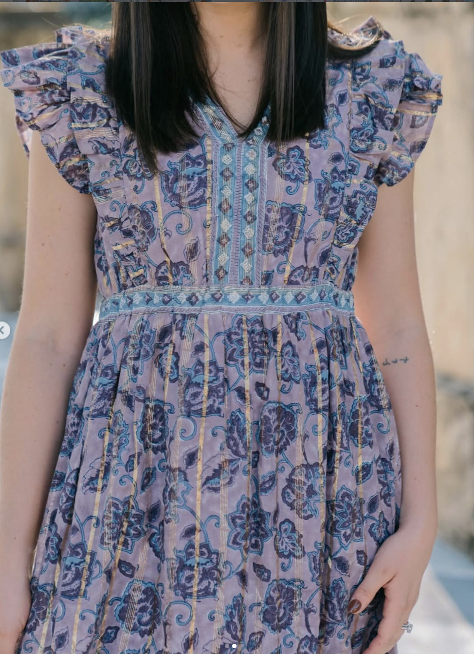 A model is wearing a floral-patterned dress with a v-neckline and short flutter sleeves, featuring intricate embroidery and a cinched waist.