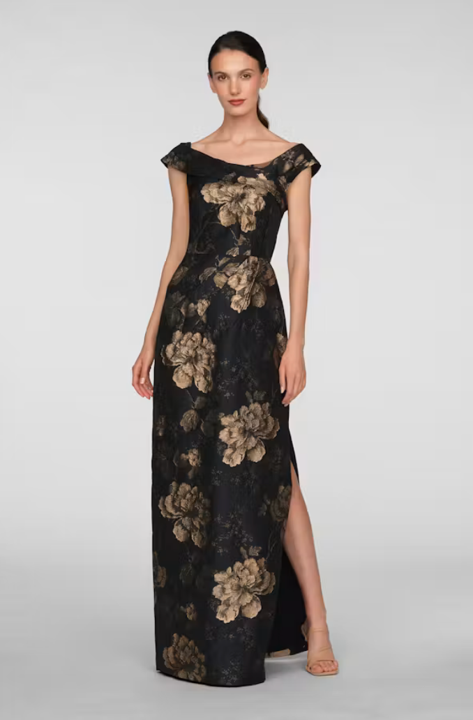A model showcases an elegant black gown adorned with large, gold floral patterns, featuring an off-the-shoulder neckline and a thigh-high slit.