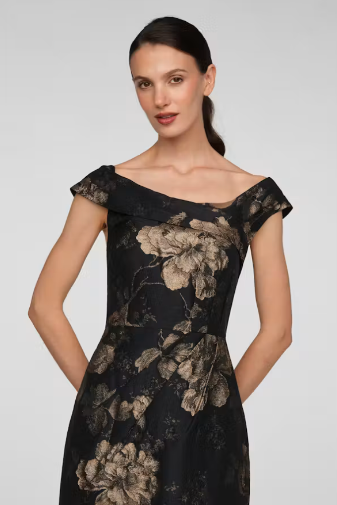 A woman models a stunning off-the-shoulder gown featuring a dark floral pattern in gold and black. This elegant dress is perfect for special occasions like a black tie wedding or gala, conveniently available near Apex, Cary, and Raleigh.