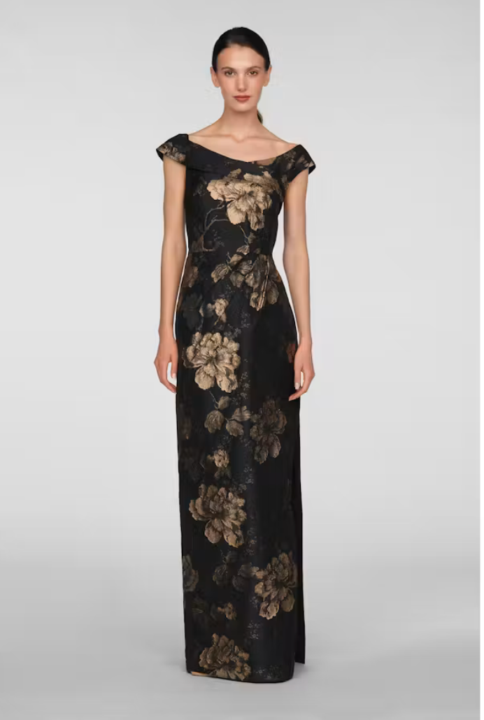 A model showcases an elegant black gown adorned with large floral patterns in gold tones, featuring an off-the-shoulder neckline and a fitted silhouette.