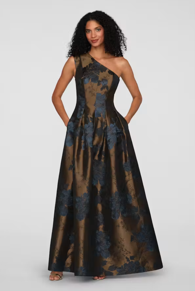 A model showcases a stunning one-shoulder gown featuring a dark floral pattern in rich tones of blue and gold.