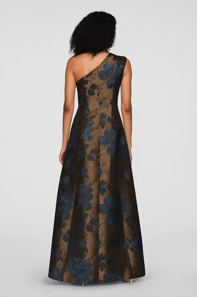 The image shows a woman wearing a one-shoulder gown featuring a floral pattern in deep blue and bronze tones.