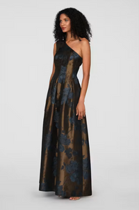 A model stands elegantly in a long, one-shoulder gown featuring a dark floral pattern. This stunning dress is perfect for formal occasions such as galas or black-tie weddings and is conveniently available near Apex, Cary, and Raleigh.