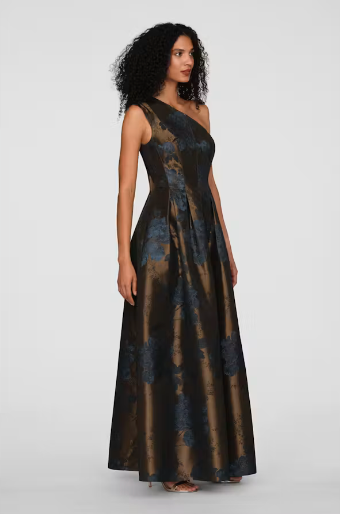 A model showcases a stunning one-shoulder gown with a rich brown and blue floral pattern, perfect for elegant occasions like black tie weddings or galas. This dress is conveniently available near Apex, Cary, and Raleigh.