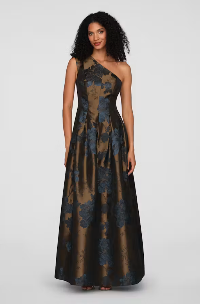 A model stands confidently in a stunning one-shoulder gown featuring a floral pattern in deep blue and gold hues.