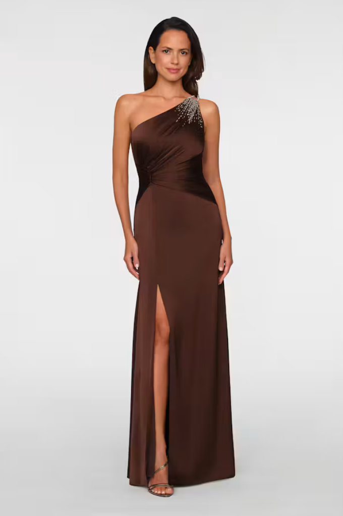 A woman models a sleek brown one-shoulder gown featuring a glamorous embellished shoulder and a thigh-high slit.
