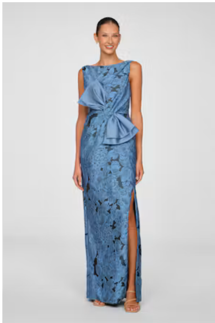 A model showcases a stunning blue gown featuring a large bow detail and a thigh-high slit, suitable for elegant occasions like a gala or black-tie wedding.
