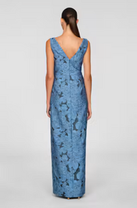 A woman stands with her back to the camera, wearing a long, elegant blue gown featuring a floral pattern.