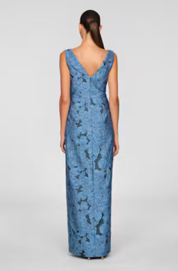 A woman stands with her back to the camera, wearing a long, elegant blue gown featuring a floral pattern.