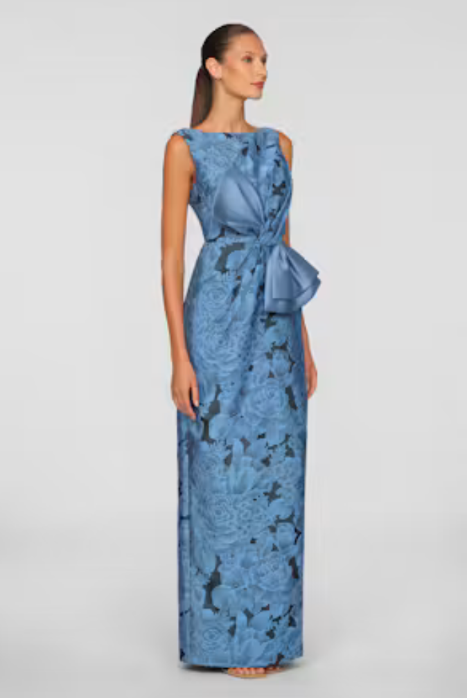 A model elegantly showcases a floor-length blue gown adorned with a floral pattern and a stylish bow at the waist.
