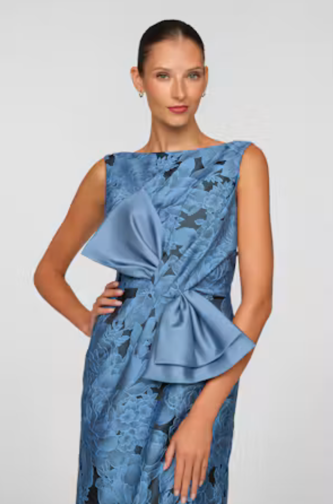 A model poses in a stunning blue gown featuring intricate floral lace and a large satin bow at the waist. This elegant dress is perfect for special events like weddings, galas, and formal occasions, available near Apex, Cary, and Raleigh.