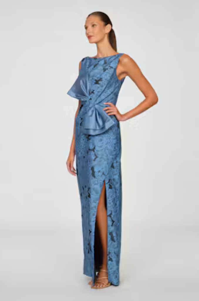 A model showcases a stunning blue gown featuring a structured bow detail at the waist and a thigh-high slit.
